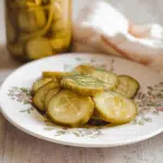 Stylish plate of crisp dill pickle slices with visible spices, next to jars of homemade pickles